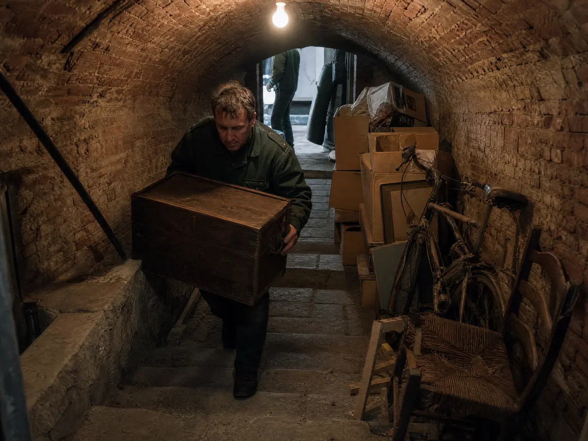 A worker carries a wooden chest up narrow stone stairs from a vaulted Brussels townhouse cellar full of boxes and an old bike